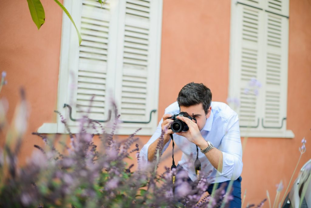 man taking aim with camera behind flower bush