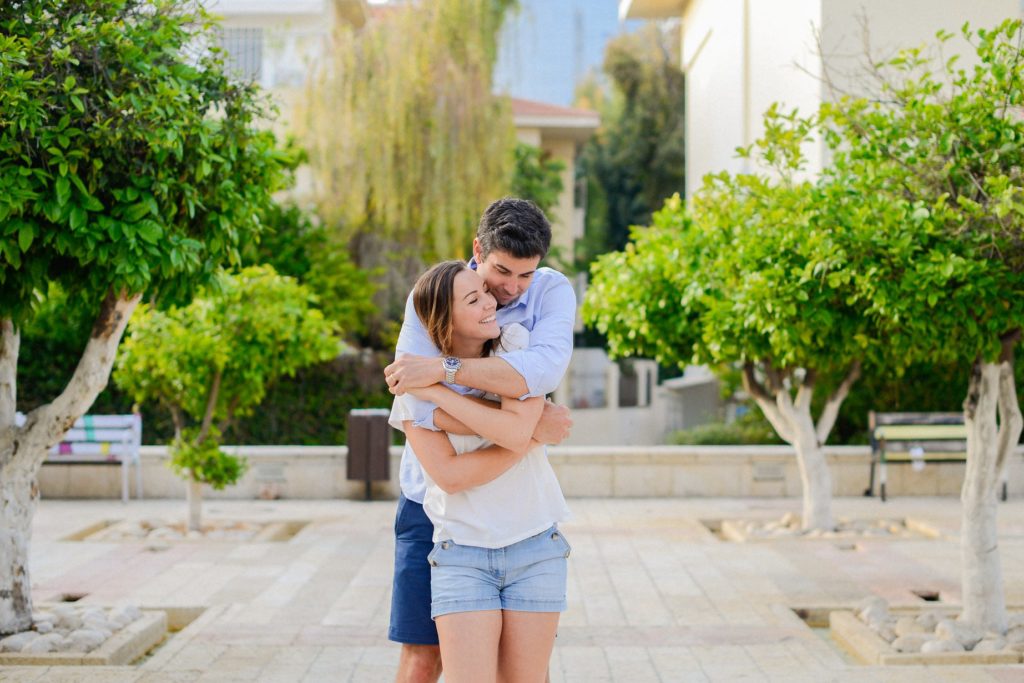 man hugging laughing woman from behind among trees in Neve Tzedek anniversary photoshoot