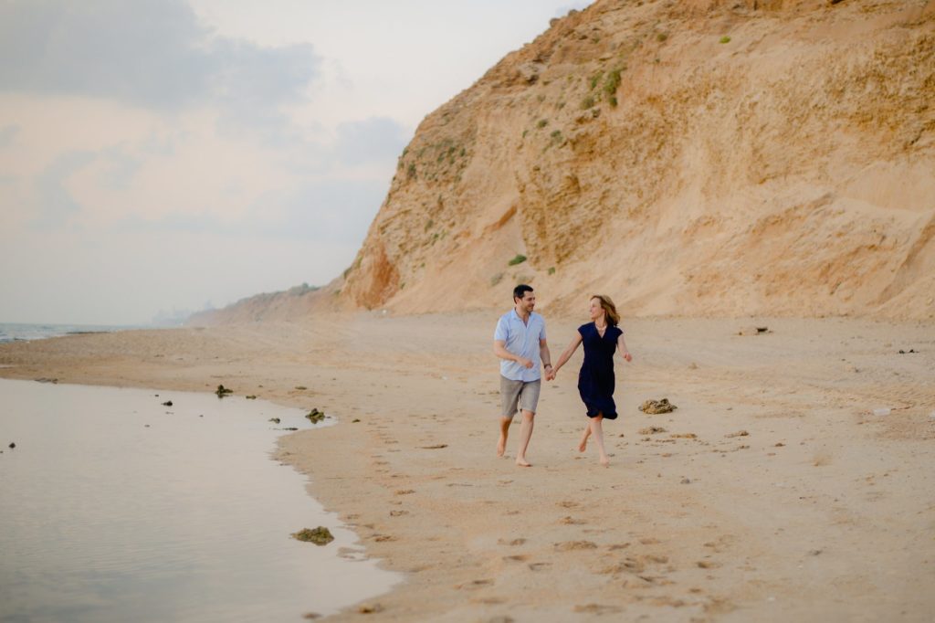 couple walking down shore and holding hands on beach engagement photoshoot