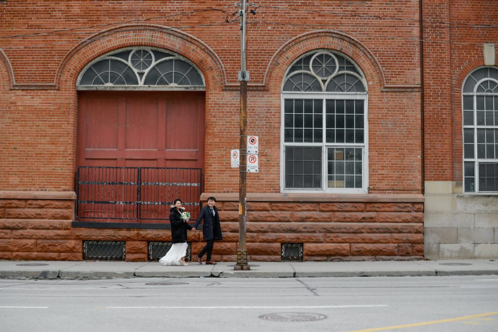 bride and groom holding hands against large red brick building for wedding photography session