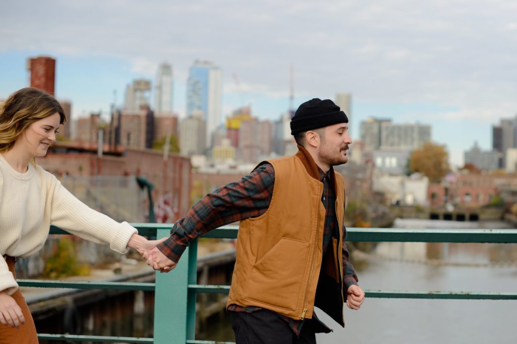 man in black beanie and brown vest pulling hand of woman in white sweater against brooklyn skyline