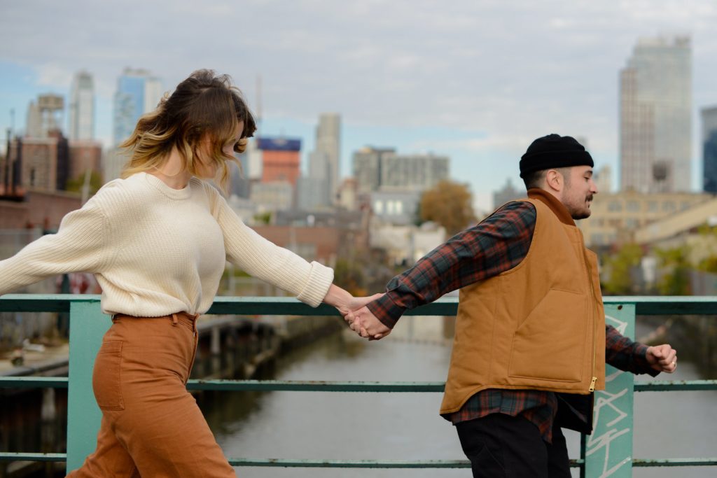 man in black beanie and brown vest holding hand of woman in white sweater, running against brooklyn skyline for engagement photoshoot