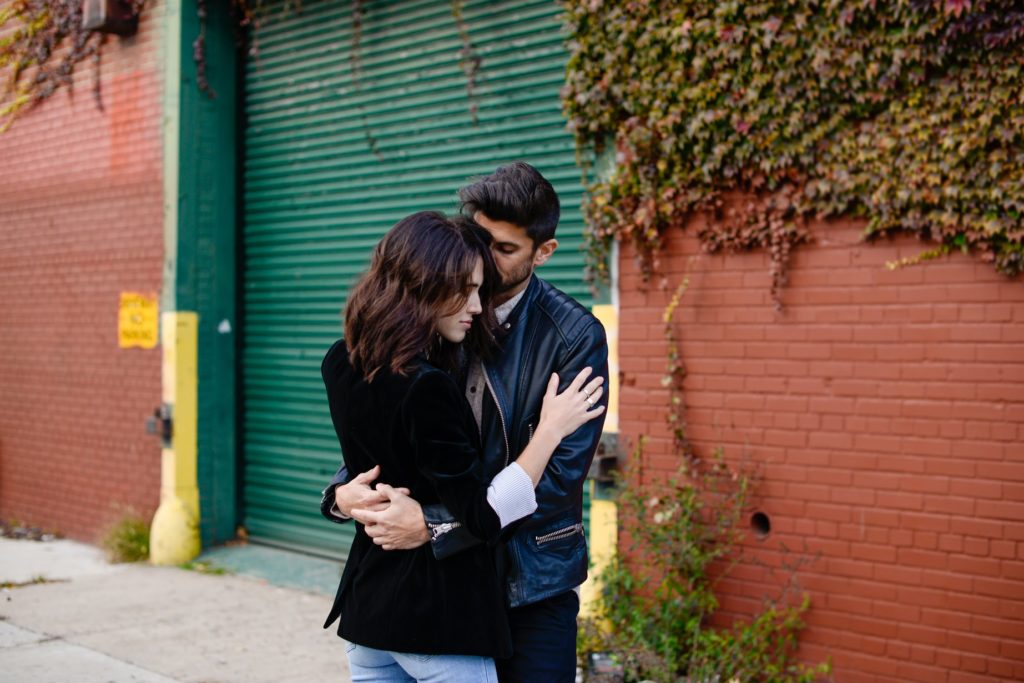 couple in black clothing hugging in front of brick wall with ivy at engagement photography session