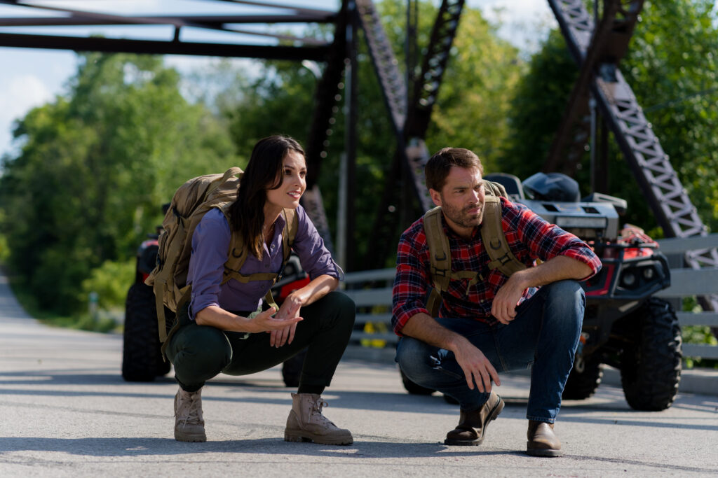 Nola Martin and Tim Rozon crouch down on a bridge to see where a lost wolf has gone in Love in Wolf Creek, a Hallmark movie